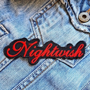 Puede incluir: Un parche negro bordado con la palabra "Nightwish" en letras rojas. El parche está cosido en una chaqueta vaquera azul, mostrando el nombre de la banda en una fuente estilizada.