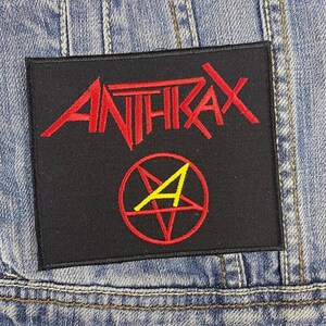 May include: A black embroidered patch featuring the word "ANTHRAX" in red, stylized lettering. Below the text is a red pentagram with a yellow "A" inside. The patch is rectangular and set against a denim background.