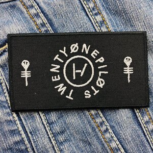 Op de afbeelding: Zwart rechthoekig embleem met wit geborduurde tekst en symbolen. De tekst luidt "TWENTY ØNE PILØTS" in een cirkel rond een centraal symbool. Twee gestileerde symbolen flankeren de tekst.