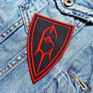 May include: A black and red embroidered patch in a shield shape. The patch features a red outline and a red stylized design on a black background. The patch is sewn onto a light blue denim jacket.
