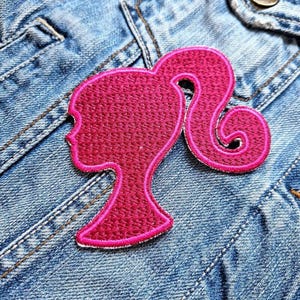 May include: A pink fabric patch in the shape of a Barbie doll silhouette. The patch is sewn onto a blue denim jacket.