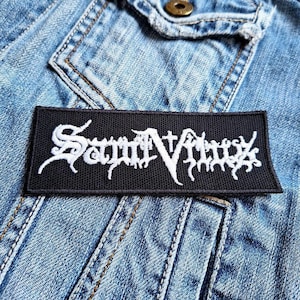 Puede incluir: Un parche negro con letras bordadas blancas que dicen "Satyricon". El parche está cosido en una chaqueta vaquera azul.