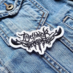 Puede incluir: Parche blanco bordado con letras negras que dice "Dethklok" en una chaqueta vaquera azul.