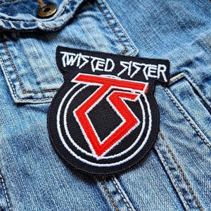 Puede incluir: Parche bordado en blanco y negro con el texto "Twisted Sister" en blanco y un logotipo estilizado "TS" en rojo y blanco. El parche está en una chaqueta vaquera azul.