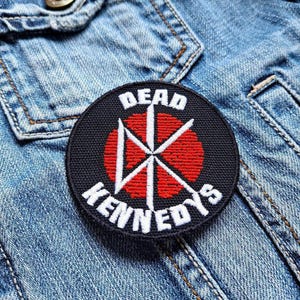 Può includere: Una toppa ricamata nera e rossa con le parole "Dead Kennedys" e un simbolo rosso e bianco.