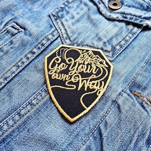 Op de afbeelding: Een geborduurde patch in zwart en goud met de tekst "Go your own way" op een spijkerjack.