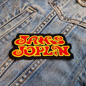 Könnte beinhalten: Ein gestickter schwarzer und roter Aufnäher mit dem Text "Janis Joplin" in einem Retro-Schriftart-Stil.