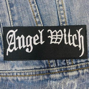 Może przedstawiać: Czarna naszywka haftowana z białym napisem "Angel Witch".