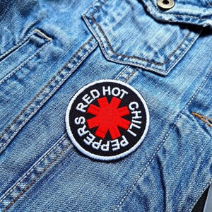 Peut inclure: Patch du logo du groupe Red Hot Chili Peppers sur une veste en jean bleue. Le patch est noir et blanc avec une étoile rouge au centre et les mots "Red Hot Chili Peppers" autour de l'étoile.