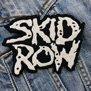 May include: Black and white embroidered patch with the text "SKID ROW" in a stylized font. The patch is on a denim background, with visible stitching and a textured appearance.