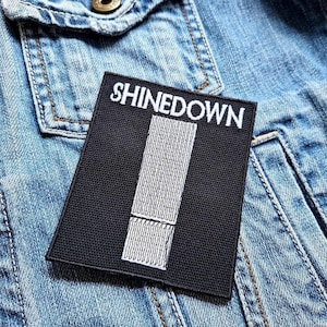 以下が含まれることがあります： 黒と白の刺繍パッチで、バンド名「Shinedown」が白文字で書かれています。パッチには、白の縦縞模様が施されています。