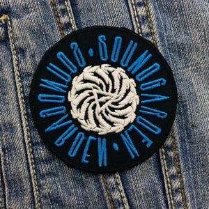 Può includere: Toppa rotonda nera con la scritta "SOUND GARDEN" in blu. Un disegno a spirale bianco è al centro. La toppa è cucita su una giacca di jeans blu, per un look classico.