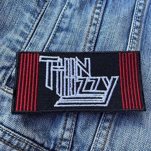 May include: Black embroidered patch with red stripes on the sides. The patch says "Thin Lizzy" in white lettering.