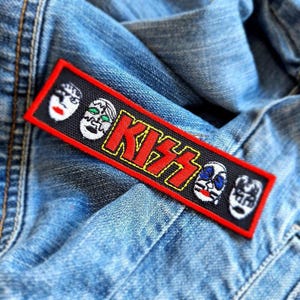 May include: A rectangular black patch with a red border featuring the word "KISS" in red and yellow lettering. The patch also displays four stylized faces in white, black, and various colors, set against a blue denim background.