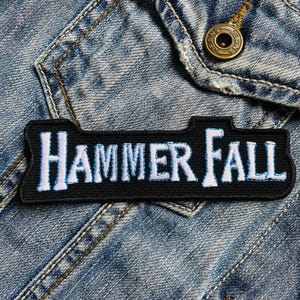 Puede incluir: Parche bordado en blanco y negro con el texto "HAMMER FALL" en letras blancas con un contorno azul. El parche está en una chaqueta vaquera azul.