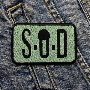 May include: A rectangular patch with the letters "S.O.D." embroidered in black on a light green background. The patch is bordered in black and is set against a denim jacket.