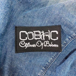 Op de afbeelding: Een zwart rechthoekig embleem met witte tekst "COBHC Children Of Bodom", genaaid op een blauw spijkerjasje. Het embleem is gecentreerd en de tekst is gestileerd.