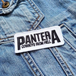 May include: A rectangular white patch with black embroidered text that reads "PANTERA COWBOYS FROM HELL." The patch is sewn onto a light blue denim jacket. The jacket has visible seams and a metal button.
