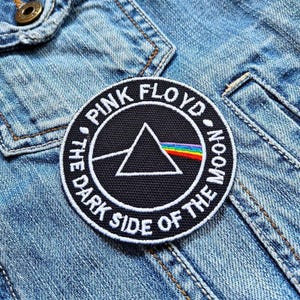 May include: A round black patch with white embroidered text and a white triangle with a rainbow prism design. The text reads "PINK FLOYD" and "THE DARK SIDE OF THE MOON". The patch is sewn onto a light blue denim jacket.