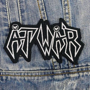 Könnte beinhalten: Schwarz-weißes Bügeleisen-Patch mit dem Wort "AT WAR" in einer stilisierten Schriftart. Das Patch hat ein Schädel-Design oben auf jedem Buchstaben.