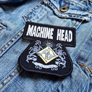 Könnte beinhalten: Schwarz-weißes Machine Head Band-Patch mit einem Wappen, das einen gelben Diamanten mit den Initialen der Band und einem Sonnenstrahl darüber zeigt. Zwei Löwen flankieren den Diamanten, und der Text "MCMXCIX" befindet sich darunter.