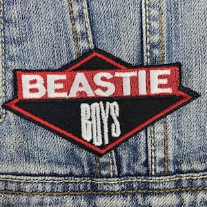 Peut inclure: Un écusson en forme de losange rouge et noir avec du texte brodé blanc qui dit "BEASTIE BOYS".