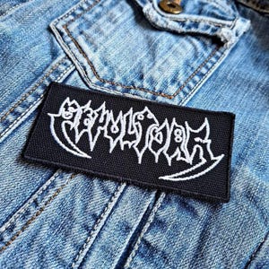Könnte beinhalten: Schwarz-weiß gestickter Patch mit dem Wort "Slaughter" in einer stilisierten Schriftart. Der Patch ist auf eine blaue Jeansjacke genäht.