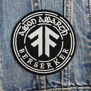 May include: A circular black and white patch with the words "AOD AMARCH BERSERKER" around the edge. The patch has a white double arrow symbol in the centre. The patch is attached to a denim jacket.