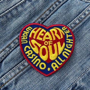 Pode incluir: Um patch em forma de coração com as palavras "HEART OF SOUL" em amarelo, vermelho e azul. O patch também inclui as palavras "CASINO ALL NIGHTER" e "WIGAN" em uma borda azul. O patch é costurado em uma jaqueta jeans.
