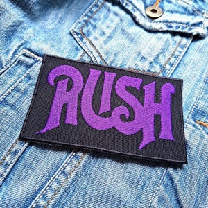 Könnte beinhalten: Lilafarbener "RUSH" Band-Logo-Patch auf schwarzem Hintergrund. Der Patch ist auf eine blaue Jeansjacke genäht.