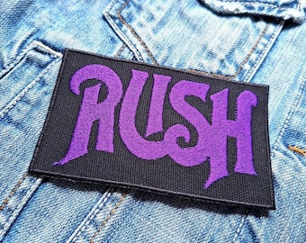 Peut inclure: Patch de logo de groupe "RUSH" violet sur fond noir. Le patch est cousu sur une veste en jean bleue.
