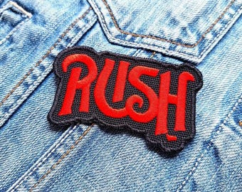 Peut inclure: Un patch brodé noir et rouge avec le mot "RUSH" dans une police de caractères stylisée.