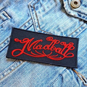 May include: A rectangular black patch with the word "Modball" embroidered in red script. The patch is affixed to a light blue denim jacket, with visible stitching and a button.