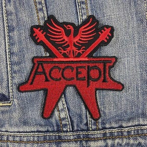 May include: A red and black embroidered patch with the word "ACCEPT" and a bird design with crossed guitars. The patch is attached to a denim jacket. The patch is a music-themed accessory.