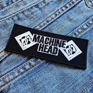 May include: Black and white embroidered patch with the text "MACHINE HEAD" and two stylised logos. The patch is on a blue denim background.