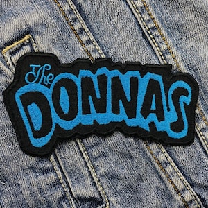 Könnte beinhalten: Ein schwarz-blauer, gestickter Aufnäher mit dem Text "The Donnas" in einer stilisierten Schriftart. Der Aufnäher ist rechteckig mit einem schwarzen Rand und vor einem Jeanshintergrund.