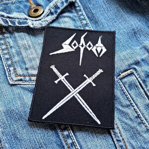 Può includere: Toppa rettangolare nera con testo e immagini ricamate in bianco. La toppa presenta la parola "Sodom" in un font stilizzato sopra due spade incrociate. La toppa è posta su una giacca di jeans blu.
