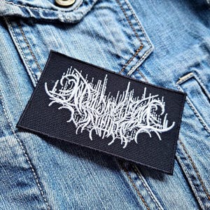 Könnte beinhalten: Ein schwarzer rechteckiger Aufnäher mit weiß gesticktem Text, möglicherweise ein Bandlogo, ist auf einer hellblauen Jeansjacke befestigt. Der Text ist stilisiert und aufwendig, mit einer gotischen oder Metal-Ästhetik.