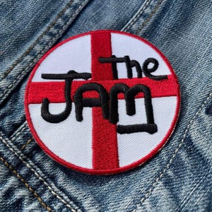 May include: A white and red circular patch with a black embroidered design of the words "The Jam" with a stylized cross in the background.
