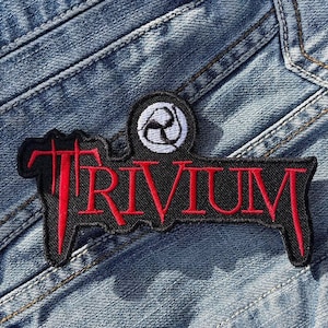 May include: Black and red embroidered patch with the word "Trivium" in red lettering. The patch has a small white circle with a black design in the upper left corner.
