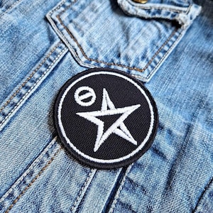 May include: Black and white embroidered patch with a star and a circle with a diagonal line through it. The patch is sewn onto a blue denim jacket.