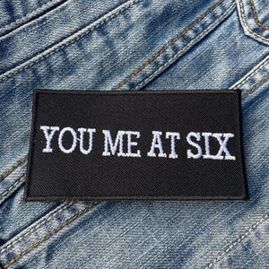 Pode incluir: Um patch retangular preto com o texto bordado branco "YOU ME AT SIX" é exibido contra um fundo de ganga azul. O patch está centrado e o texto está em negrito, com uma fonte sem serifa.