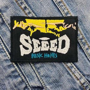 Op de afbeelding: Zwarte geborduurde patch met het woord "SEED" in wit met een geel en roze ontwerp erboven en de woorden "MUSIC HANKS" in blauw eronder.