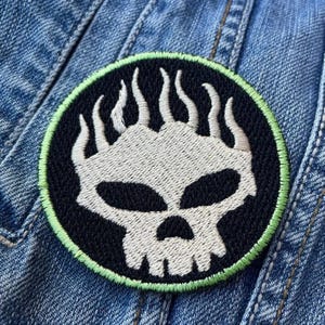 Op de afbeelding: Geborduurde patch met een schedel- en vlamontwerp op een zwarte achtergrond. De schedel en vlammen zijn wit, met een groene rand. De patch is geplaatst op een denim achtergrond.