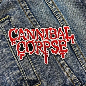 Puede incluir: Un parche bordado rojo y blanco con las palabras "CANNIBAL CORPSE" en una fuente estilizada. El parche está sobre una chaqueta vaquera azul. El texto es rojo con contorno blanco y relleno negro.
