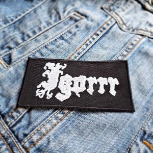 May include: Black and white embroidered patch with the word "SORRR" in a stylized font. The patch features a small, stylized creature to the left of the word.