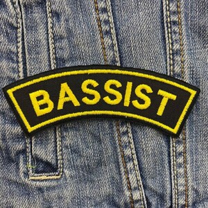 Op de afbeelding: Een zwart embleem met gele geborduurde tekst "BASSIST". Het embleem heeft een gele rand en is op een spijkerjas genaaid. Het embleem is gebogen en rechthoekig.