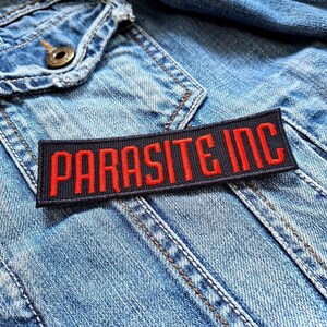 May include: A rectangular black patch with the words "PARASITE INC" embroidered in red. The patch is sewn onto a light blue denim jacket, with visible stitching and a buttoned pocket.