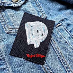 Op de afbeelding: Een zwarte patch met een witte geborduurde letter 'D' en de tekst 'Roger Waters' in rood.