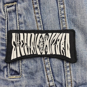 Puede incluir: Parche bordado en blanco y negro con forma rectangular. El parche presenta un diseño estilizado con la palabra "SUNRISE" en un patrón ondulado, similar a una cebra. El parche está cosido a una chaqueta vaquera.